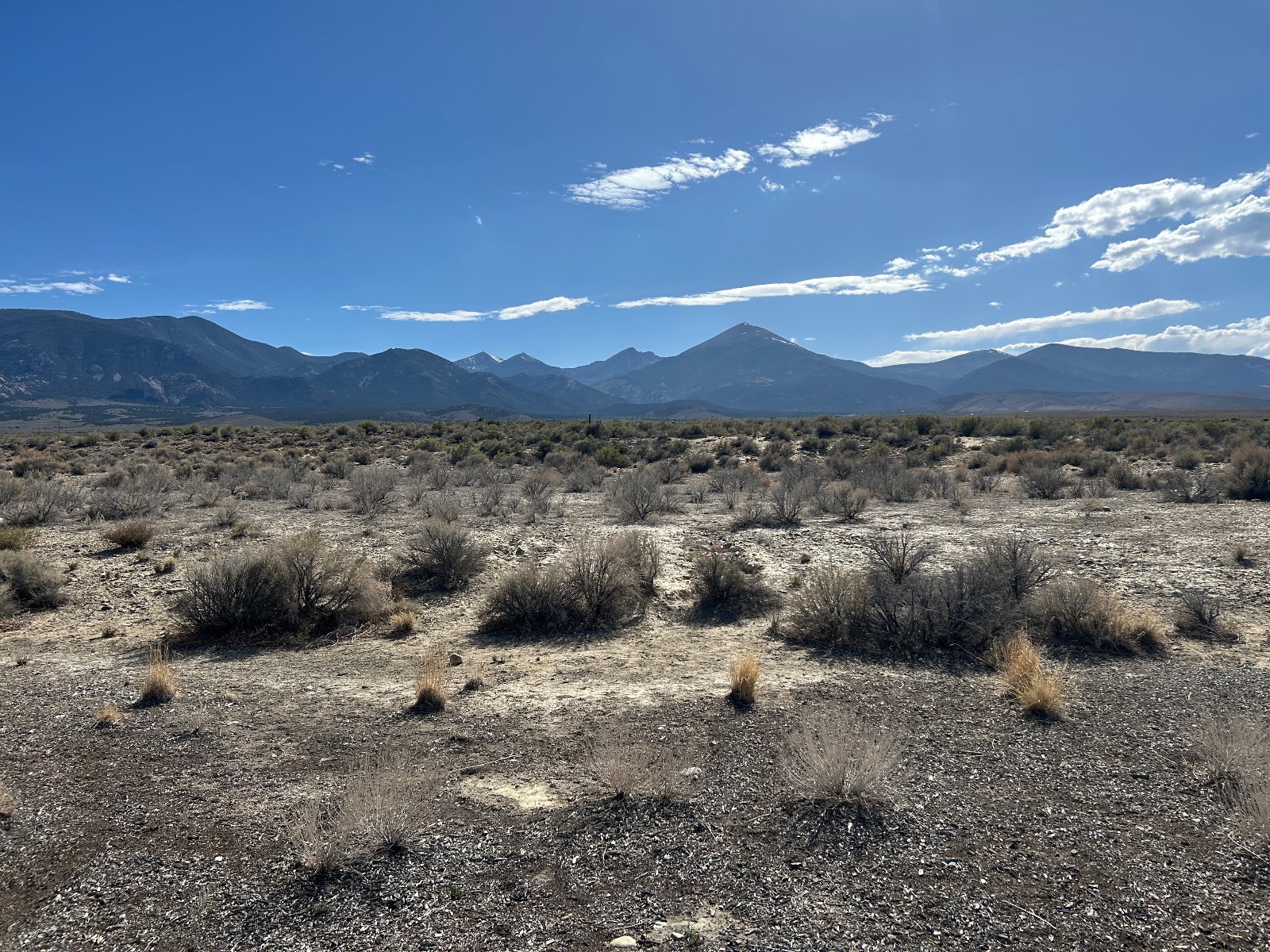 Expansive high desert landscape with sparse vegetation, gravel terrain, and snow-capped volcanic peaks in the distance under blue sky with white clouds