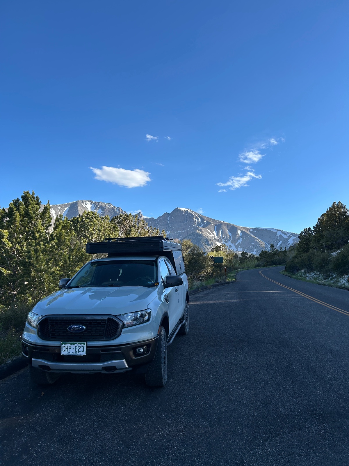 White Ford Ranger with roof rack parked on a paved mountain road with snow-capped peaks in the background under blue sky