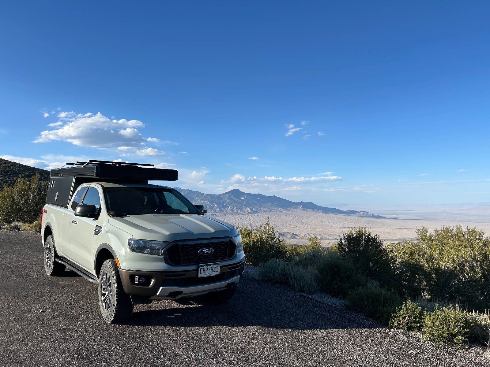 White Ford Ranger with roof rack parked at high desert overlook with mountain range and valley views under blue sky