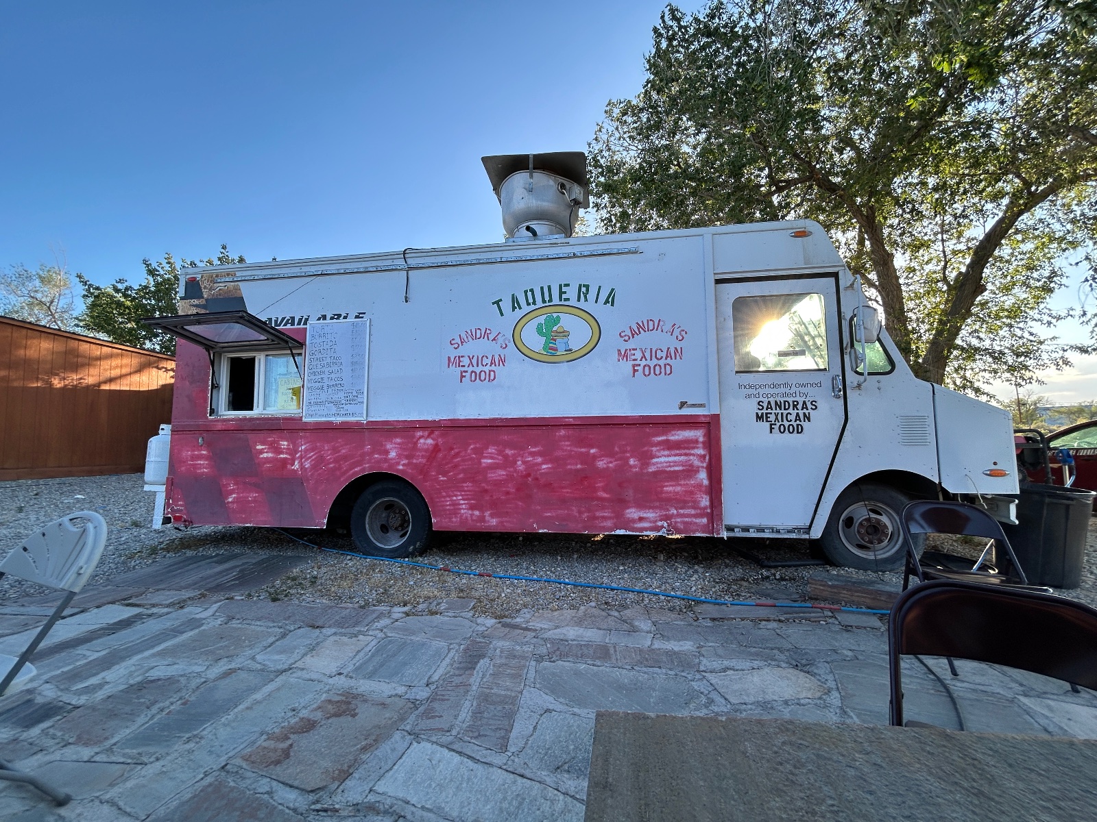 Sandra's Taqueria food truck with white and red exterior parked on stone patio under shade trees on clear day