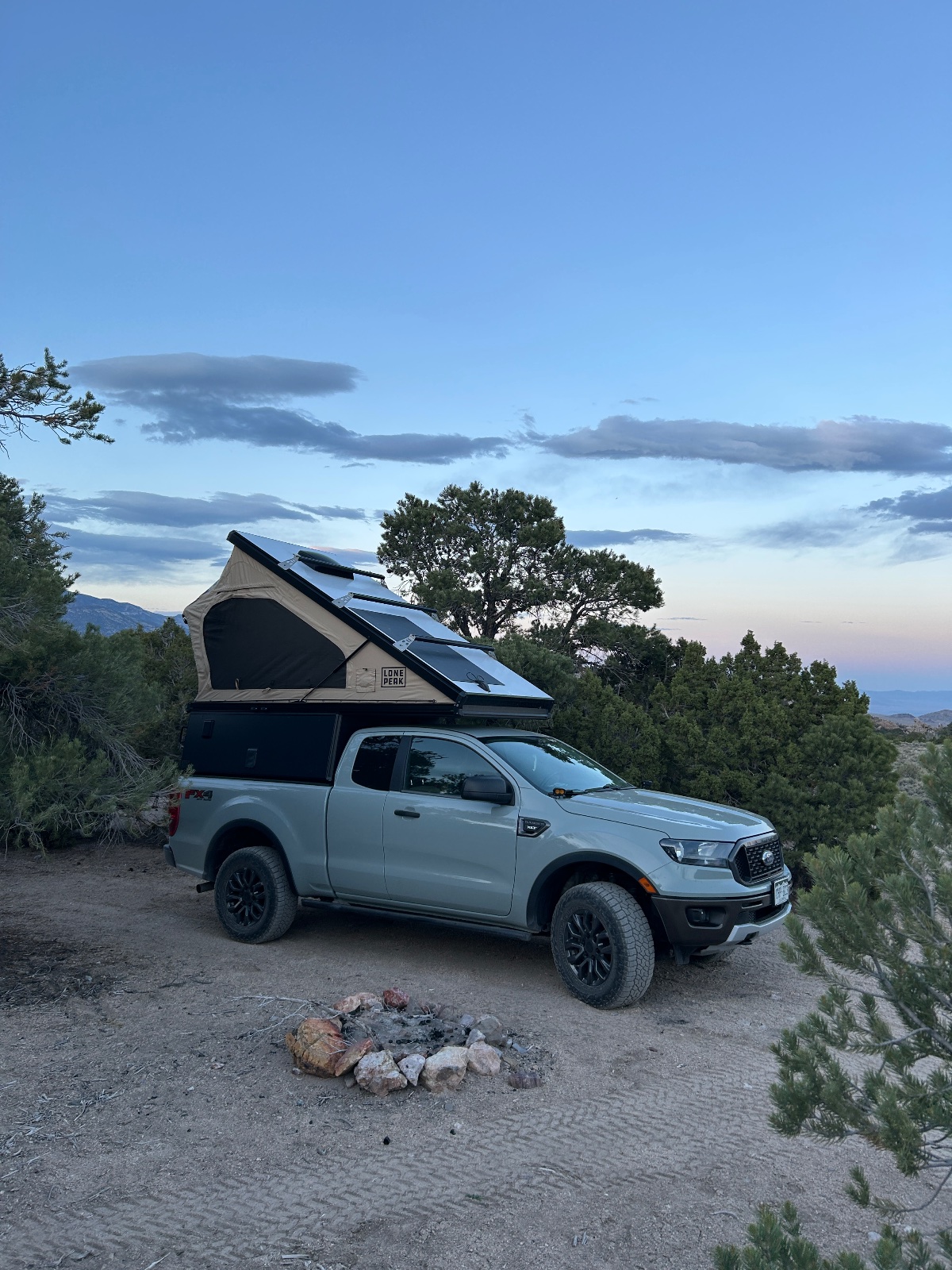 Silver Ford Ranger pickup truck with roof-mounted tent set up at a desert campsite surrounded by juniper trees and mountains at dusk