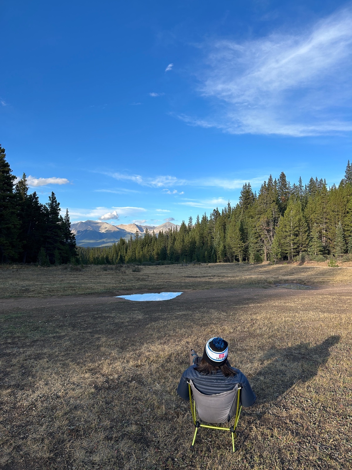 Person sitting in a camping chair overlooking a mountain lake surrounded by coniferous forest under blue sky
