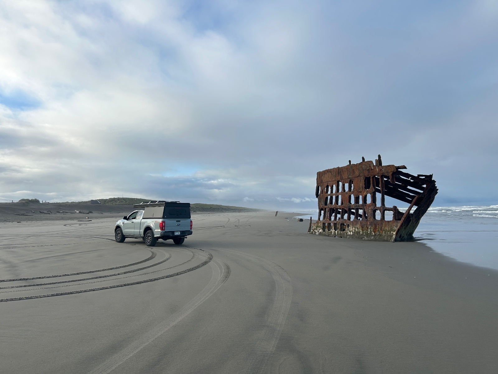 White pickup truck parked on sandy beach next to rusted shipwreck skeleton with broken hull and wooden framework, overcast sky and dunes in background