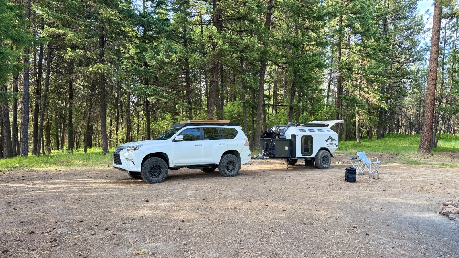 White Lexus GX460 SUV parked next to a white Mammoth off-road trailer at a forest campsite with tall pines and camping chairs visible.