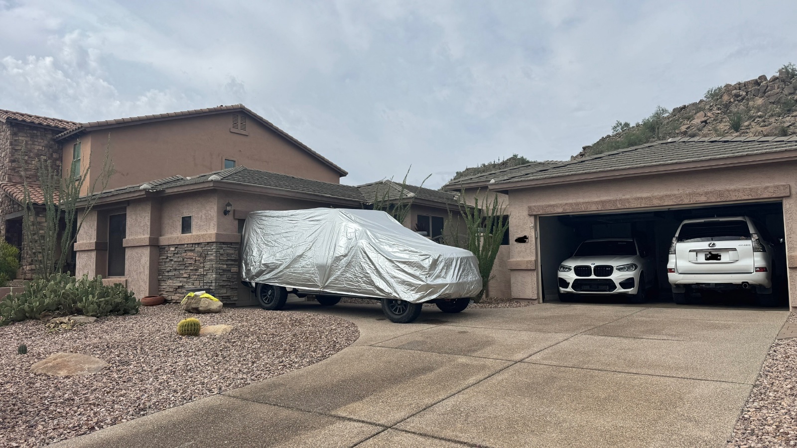 Modern home with covered BMW M car and white Lexus in garage, desert landscape with cacti and rocky hillside backdrop