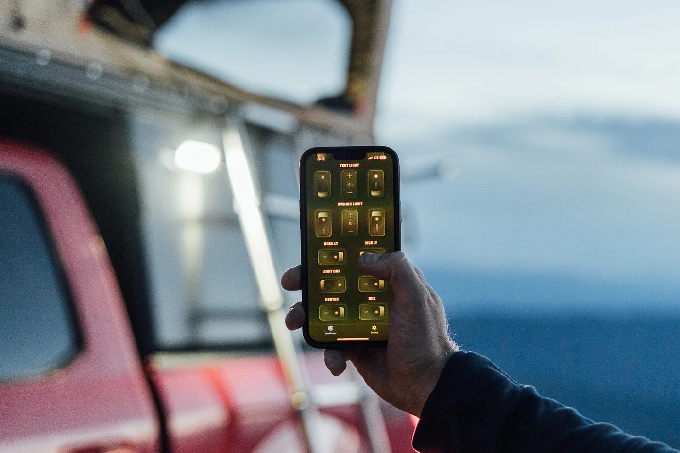 Person holding a smartphone displaying an RV electrical control panel app with various light and system controls, standing outside a red vehicle overlooking ocean water