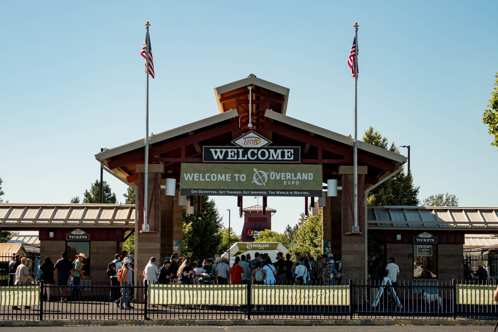 Overland Expo entrance with welcome signage, American flags, ticket booths, and visitors gathering at the gated entrance on a clear day