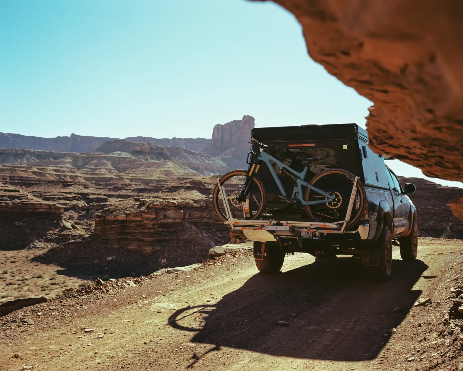 A vehicle with a bike rack carrying a mountain bike is parked on a desert trail, with red rock formations and canyon landscape visible in the background under clear blue sky.