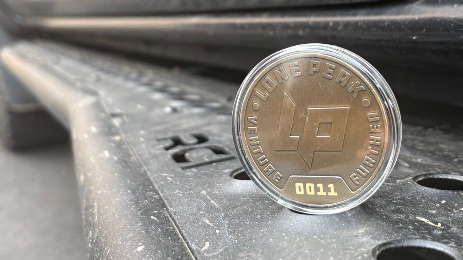 A commemorative coin in a protective case resting on weathered metal vehicle surface, featuring geometric design and numbered 0011