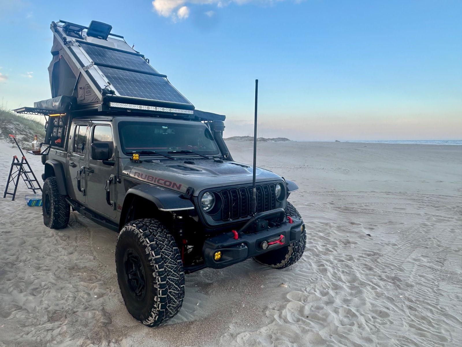 Modified Jeep Wrangler Rubicon with roof-mounted solar panels and camping gear parked on a sandy beach at the ocean's edge