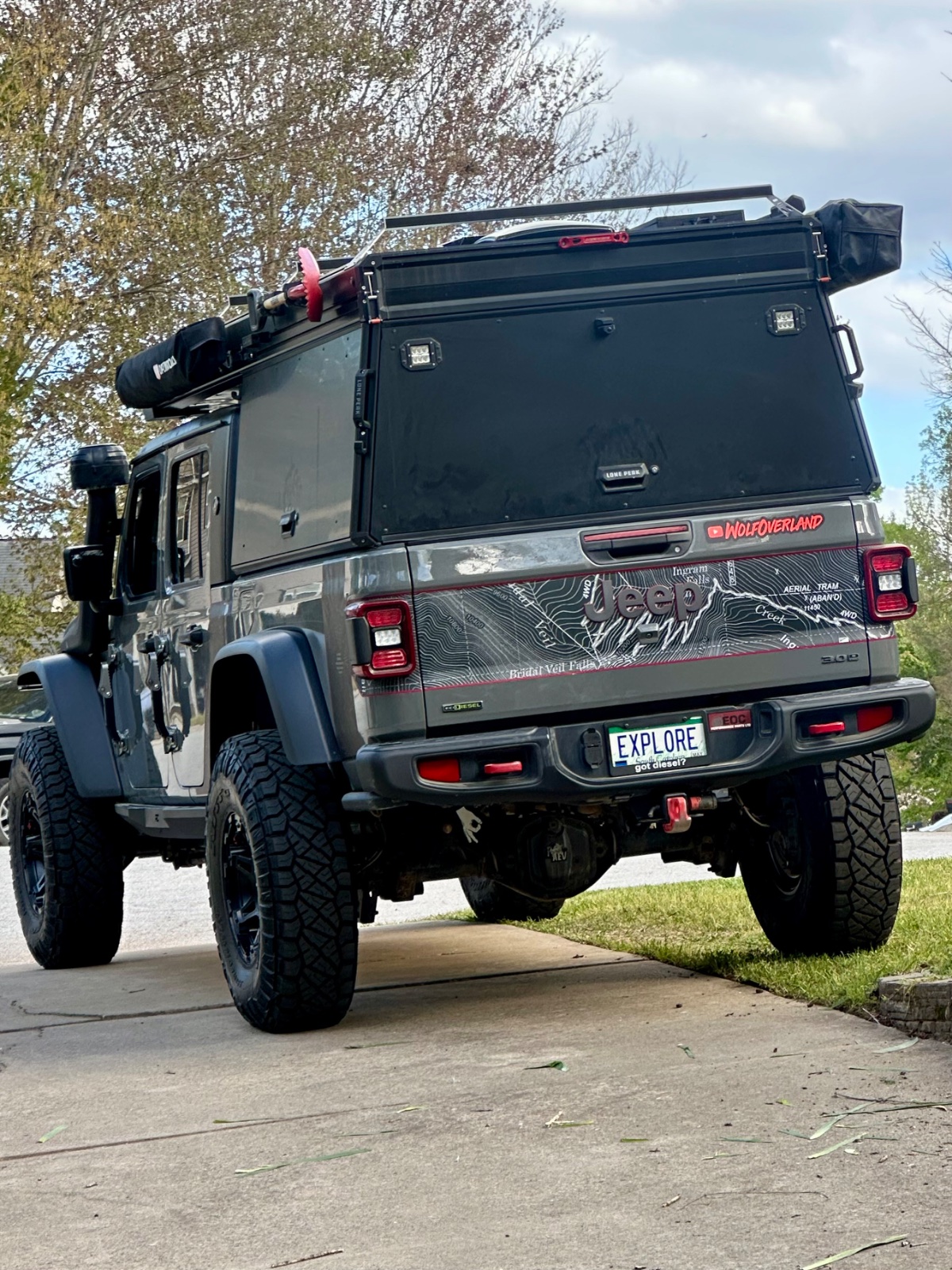 Modified Land Rover Defender 110 with roof rack, winch, and black hardtop camper shell parked on residential street with trees