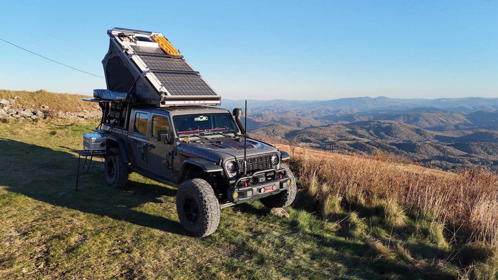 Modified Jeep Wrangler with rooftop solar panel and pop-up tent camper parked on mountainous overlook with expansive valley landscape view