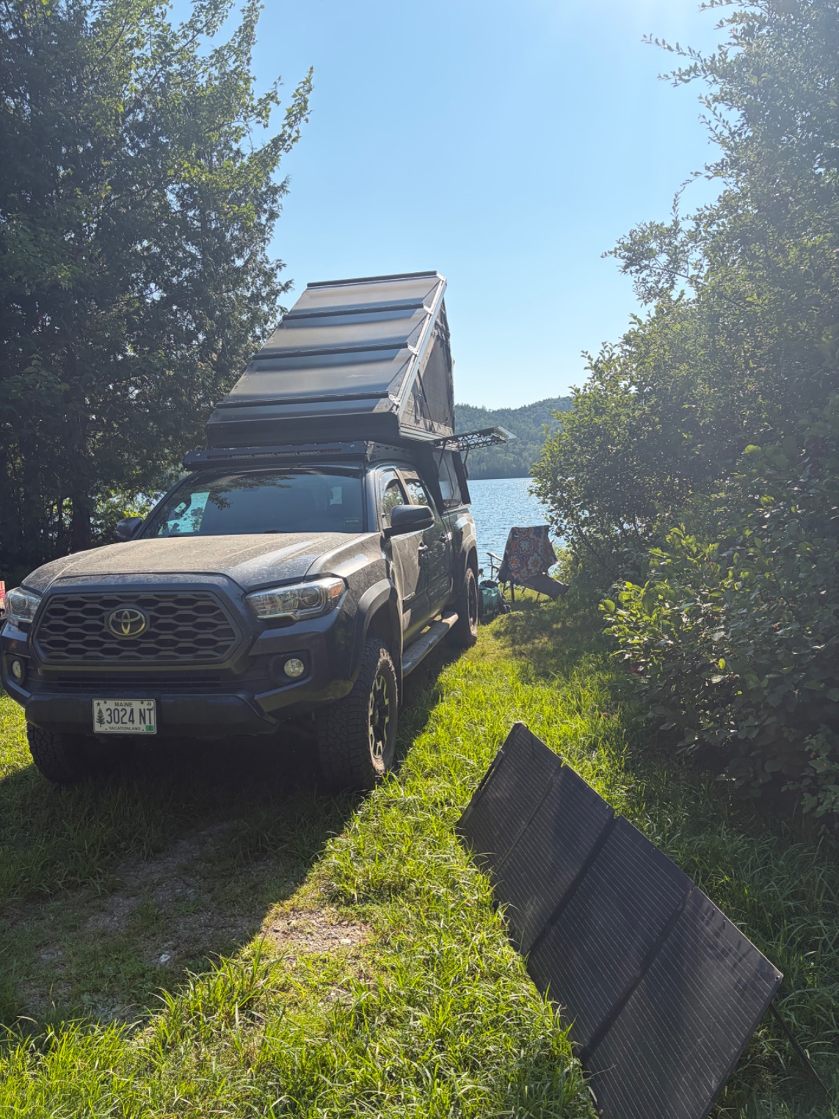 Toyota Tacoma with roof-mounted camper box parked at a lakeside camping spot surrounded by dense forest and vegetation
