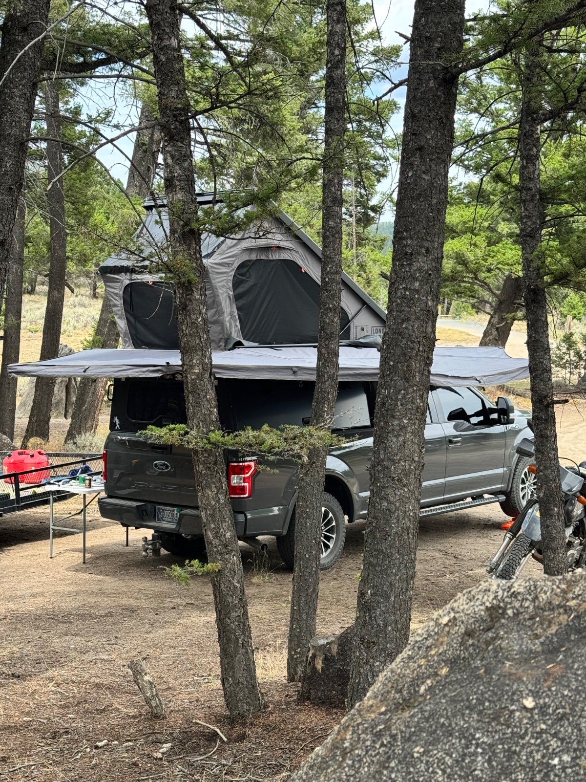 Ford F-150 pickup truck with roof-mounted tent set up in a forested campground surrounded by tall pine trees, with camping chairs and gear visible
