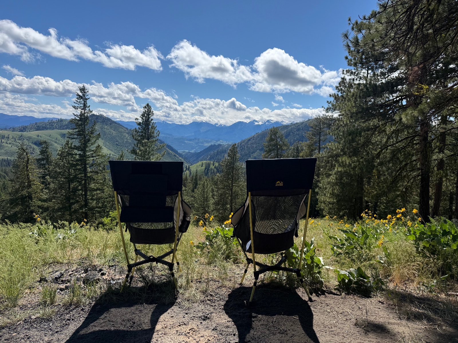 Two black camping chairs positioned on a mountain overlook with forested valleys, snow-capped peaks, and blue sky with white clouds in the distance
