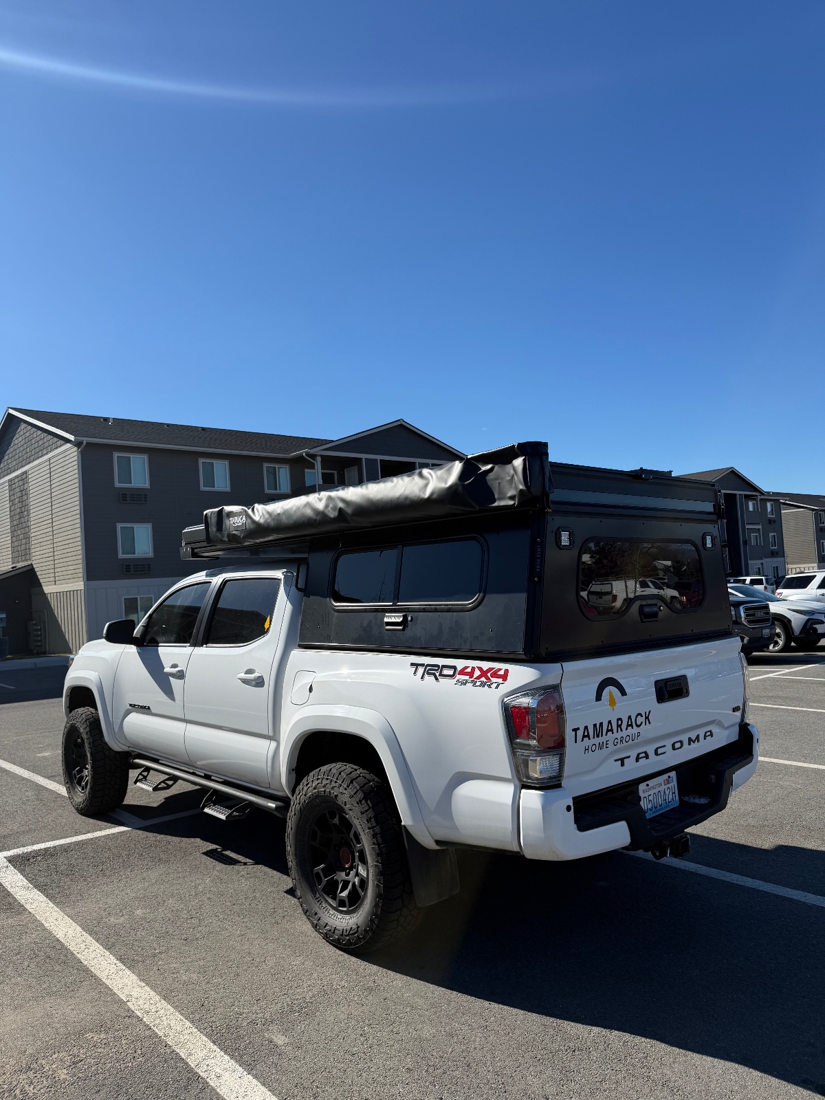 White Toyota Tacoma TRD 4x4 with black roof camper shell and rolled-up awning parked in a clear lot