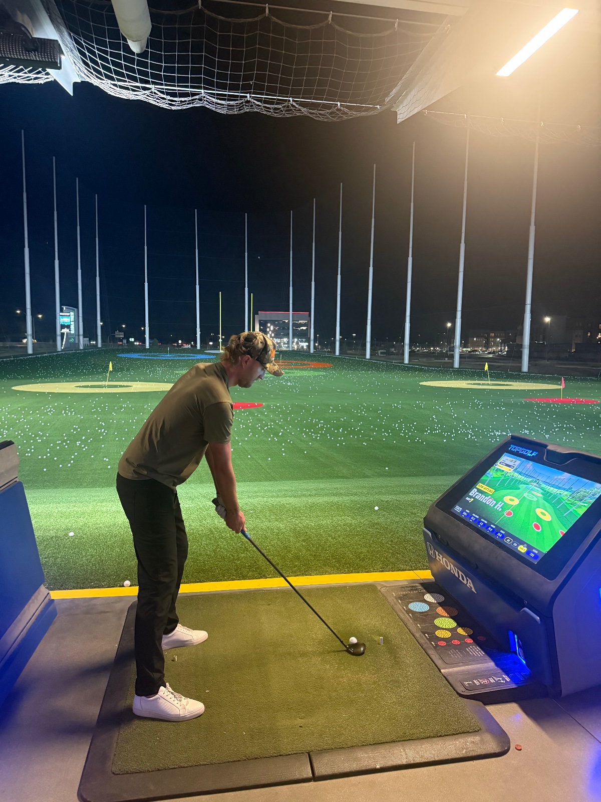 Person playing indoor simulator golf at night under bright lights with hitting mat and screen display showing course layout