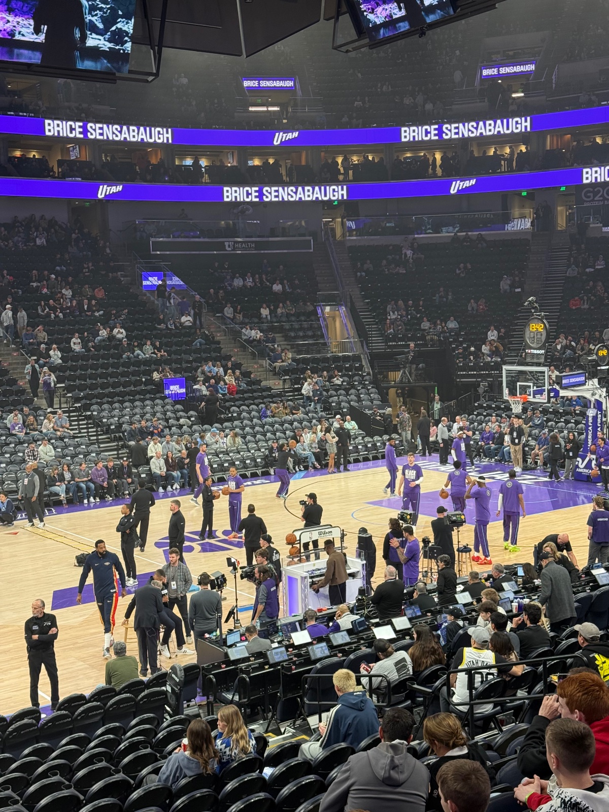 Utah Jazz basketball arena during pre-game warmup with purple court, players in uniforms, media personnel, and spectators in seats