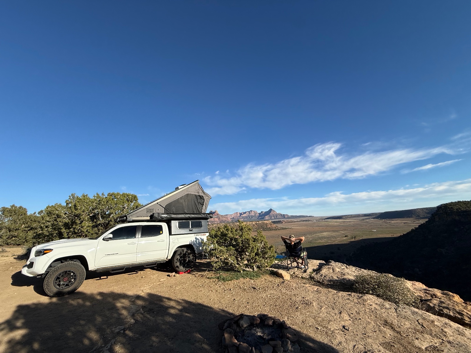 White Toyota Tacoma pickup truck with roof tent parked at a desert overlook, with person relaxing in camp chair and red rock formations visible in the distance under clear blue sky