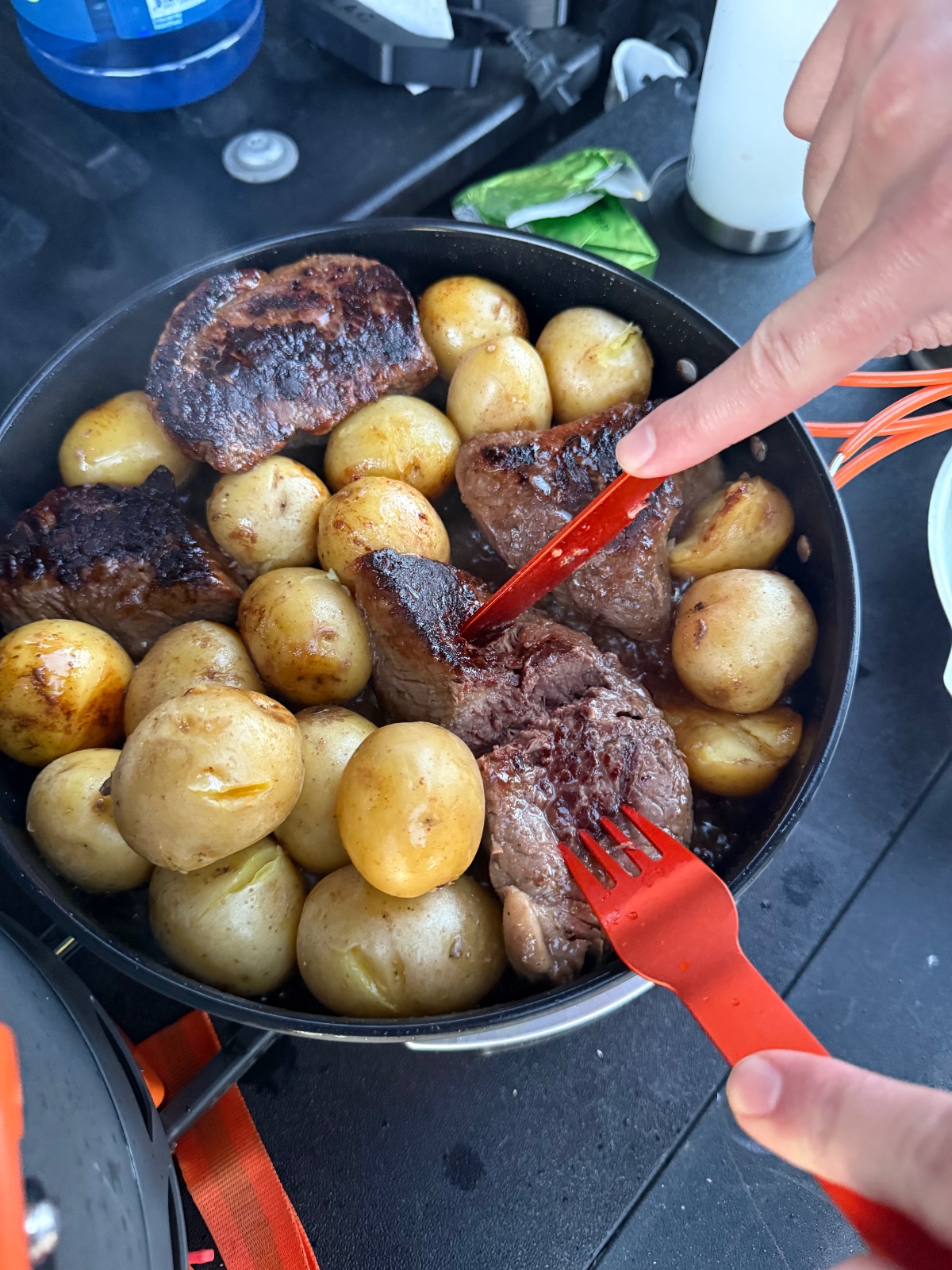 Seared steak and roasted potatoes cooking in a cast iron skillet over a portable camping stove during an outdoor adventure