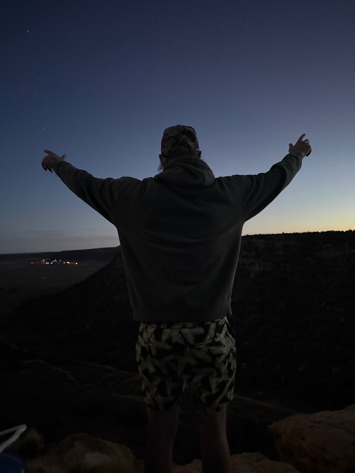 Person with arms raised standing on a rocky overlook at dusk with a scenic valley and distant lights visible below