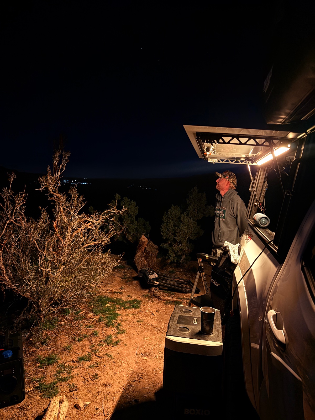 Person standing on an RV awning at night overlooking a desert landscape with sparse vegetation and distant lights