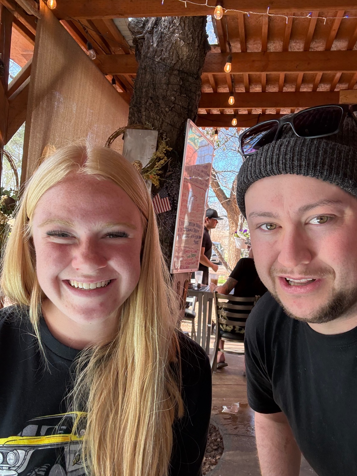 Two smiling people posing together under a wooden pergola with a large tree trunk, wearing casual outdoor clothing and sunglasses on a sunny day