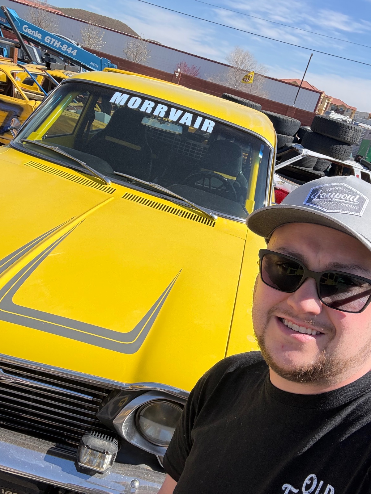 Man in sunglasses posing next to a bright yellow vintage car with 'MORRAIR' text on windshield at an outdoor vehicle meet