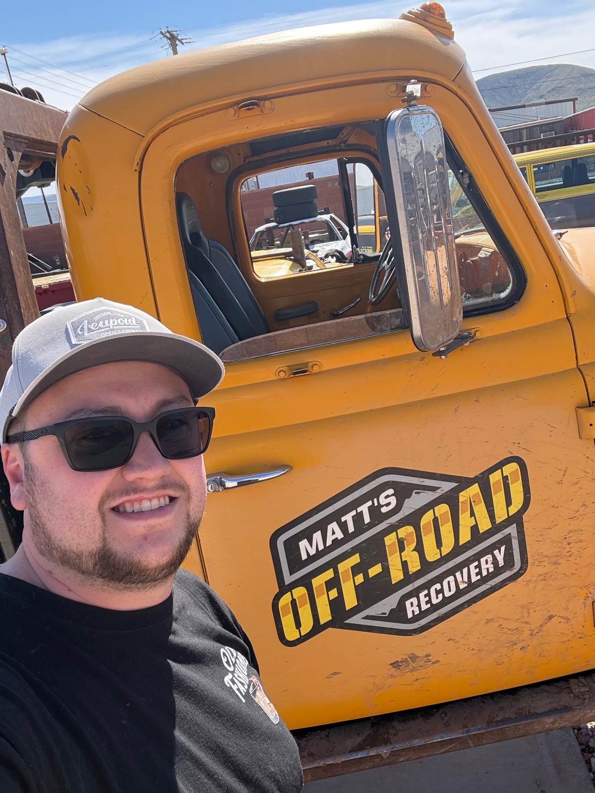 Man in cap and sunglasses poses next to a bright yellow vintage truck with 'Matt's Off-Road Recovery' branding at a desert rally or gathering