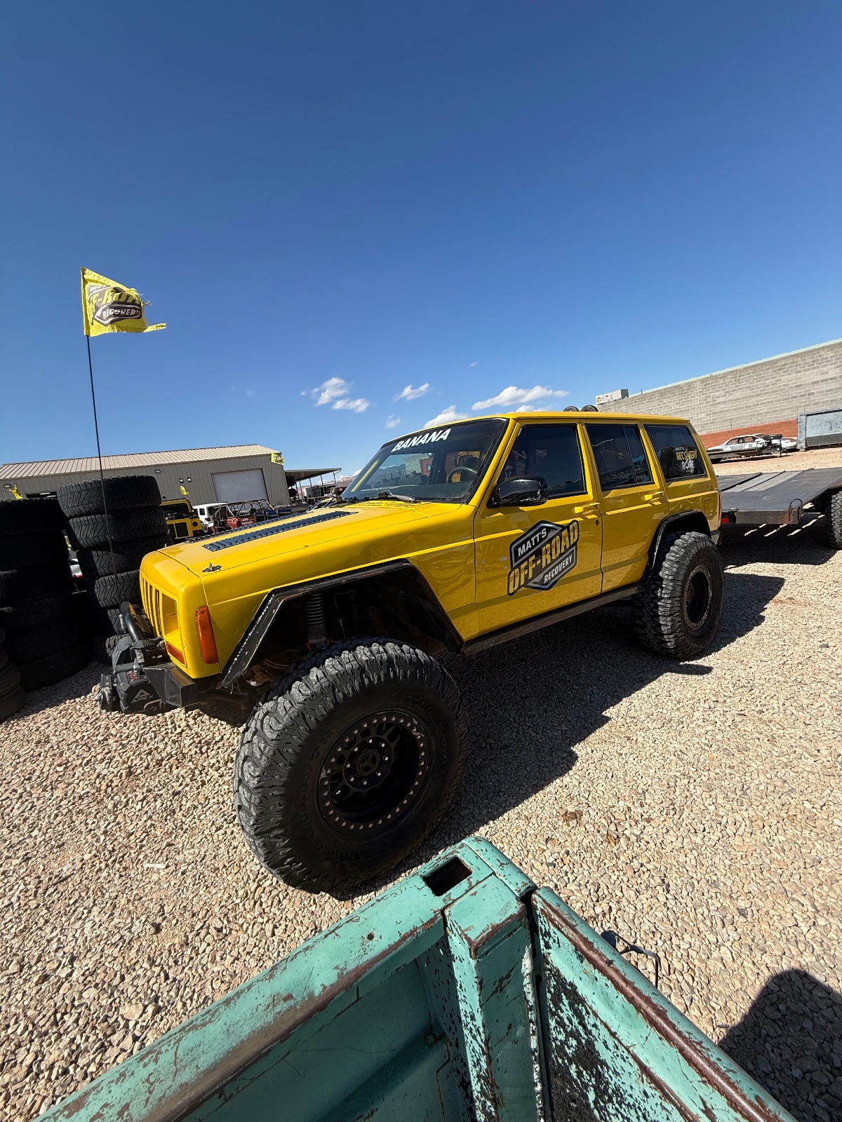 Bright yellow lifted Jeep Cherokee with oversized all-terrain tires parked at an off-road rally or event, with industrial buildings and clear blue sky in background