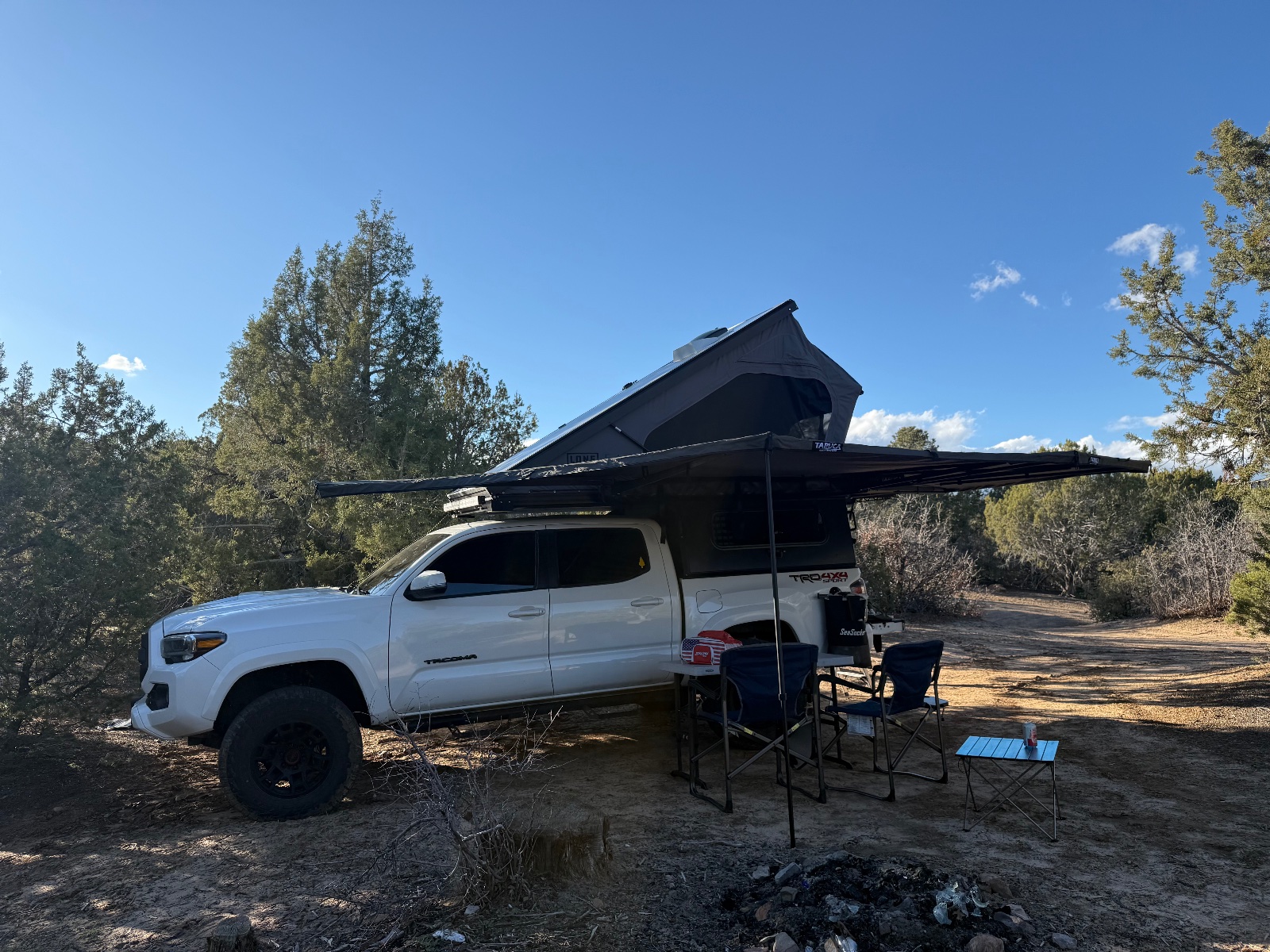 White Toyota Tacoma with roof-mounted tent and awning deployed at a desert campsite surrounded by sparse vegetation and pine trees under clear blue sky