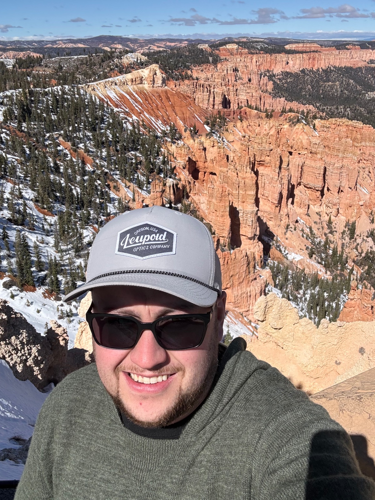 Man wearing Leupold Optics cap and sunglasses smiles at Bryce Canyon National Park rim with distinctive red hoodoo formations and snow-covered pine forest in background