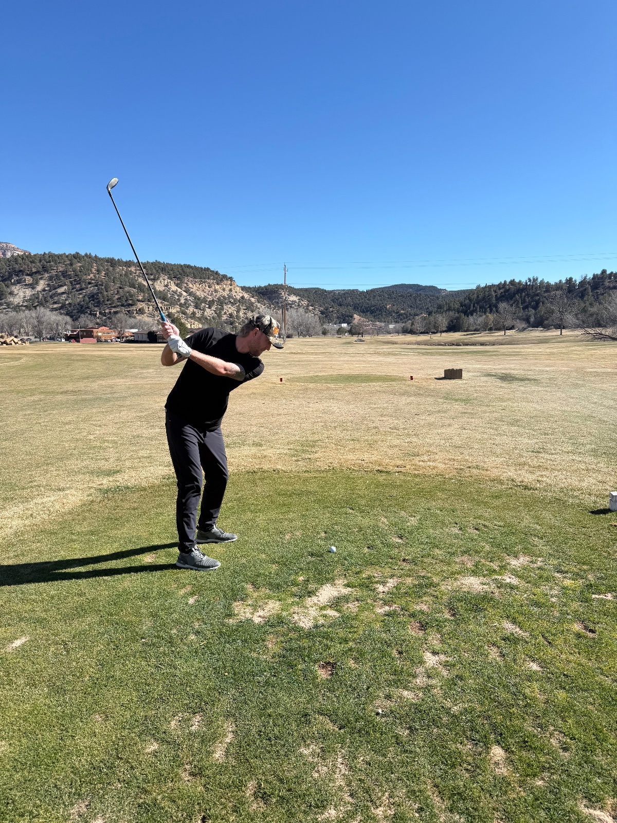Golfer mid-swing on a wide open fairway with forested mountains and clear blue sky in background at a mountain valley golf course