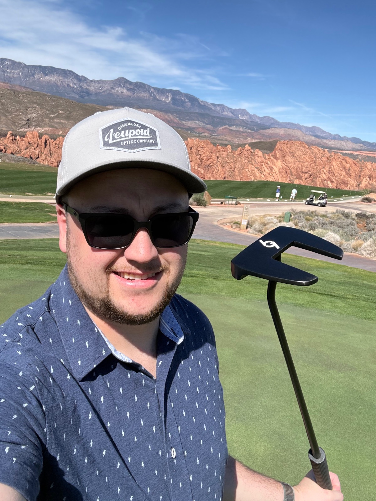 Man in Leupold Optics cap golfing at a scenic desert course with red rock formations and mountains in background