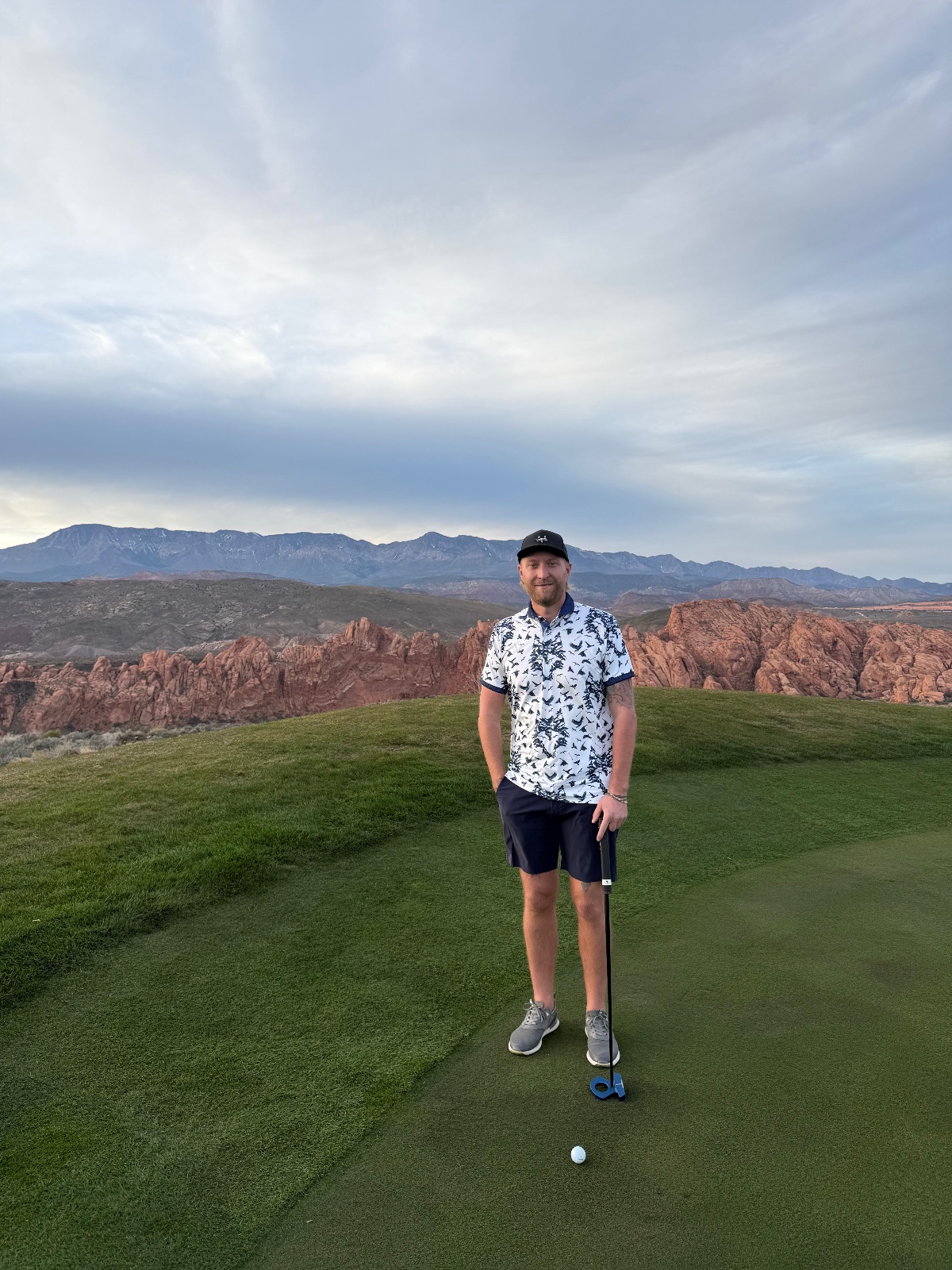 Man holding putter on manicured golf green overlooking red rock formations and distant mountains under cloudy sky