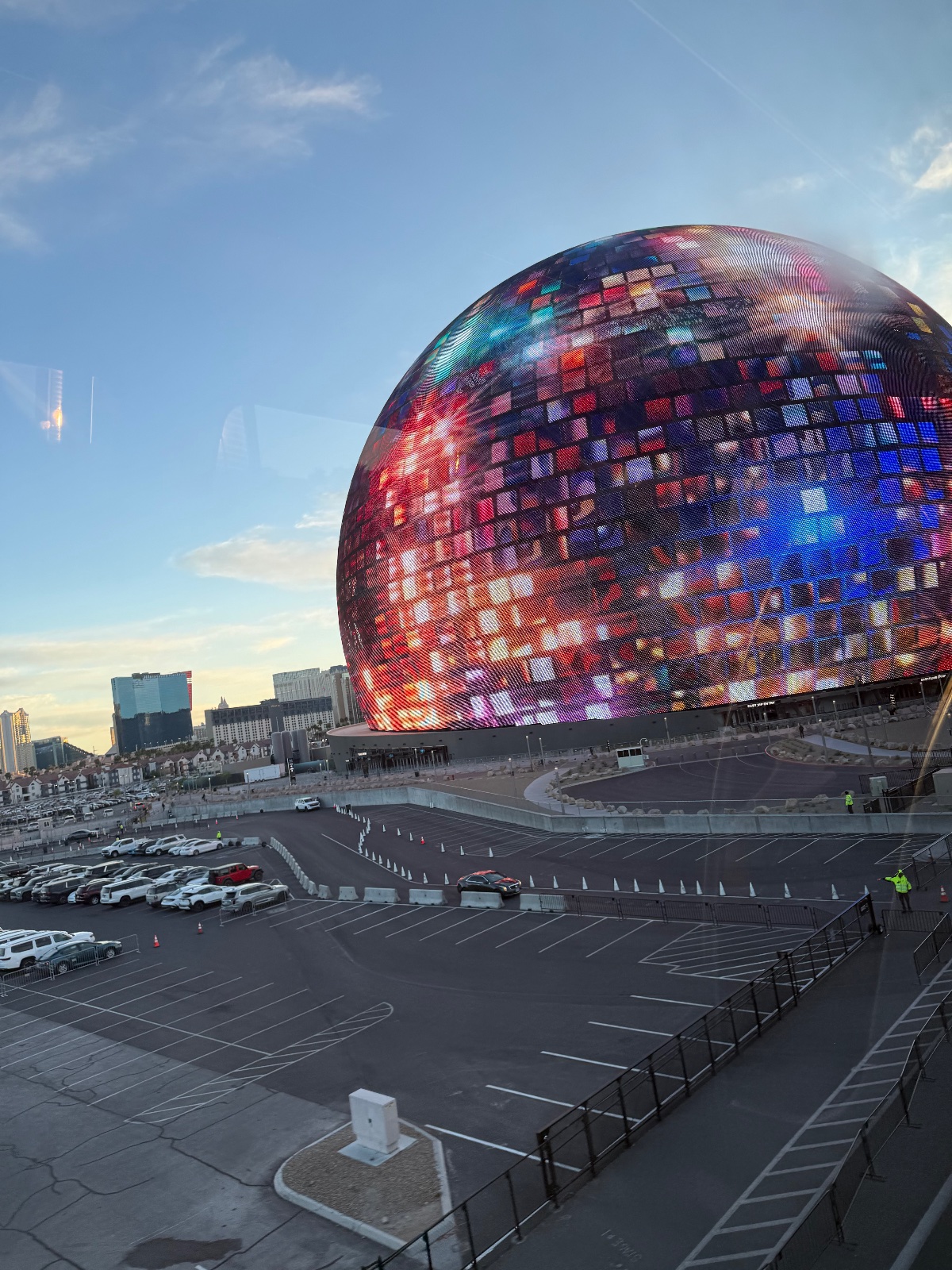 Massive illuminated spherical dome structure with colorful LED display in urban parking lot with city skyline under blue sky