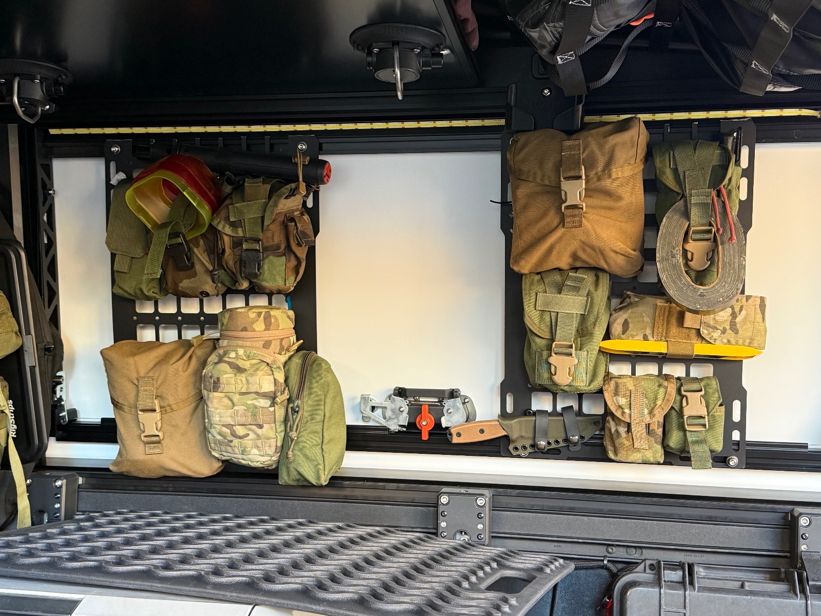 Vehicle storage wall displaying tactical gear including camouflage pouches, tan ammunition cans, coiled rope, yellow pry bar, and various olive drab MOLLE-style bags organized on black mounting rails.