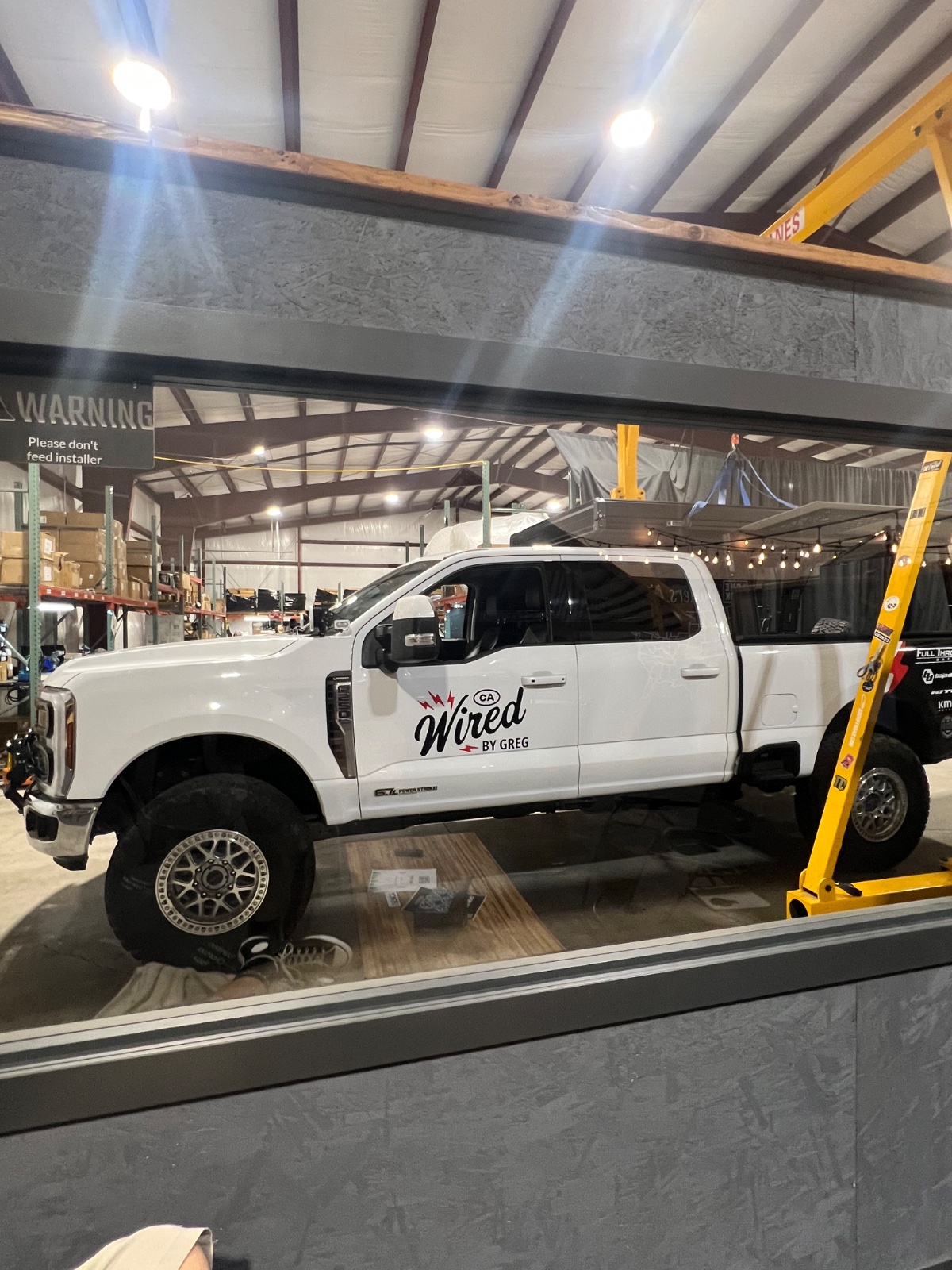 White Ford F-250 Super Duty truck with 'Wired by Greg' branding parked on a lift in an industrial workshop with yellow lifting equipment