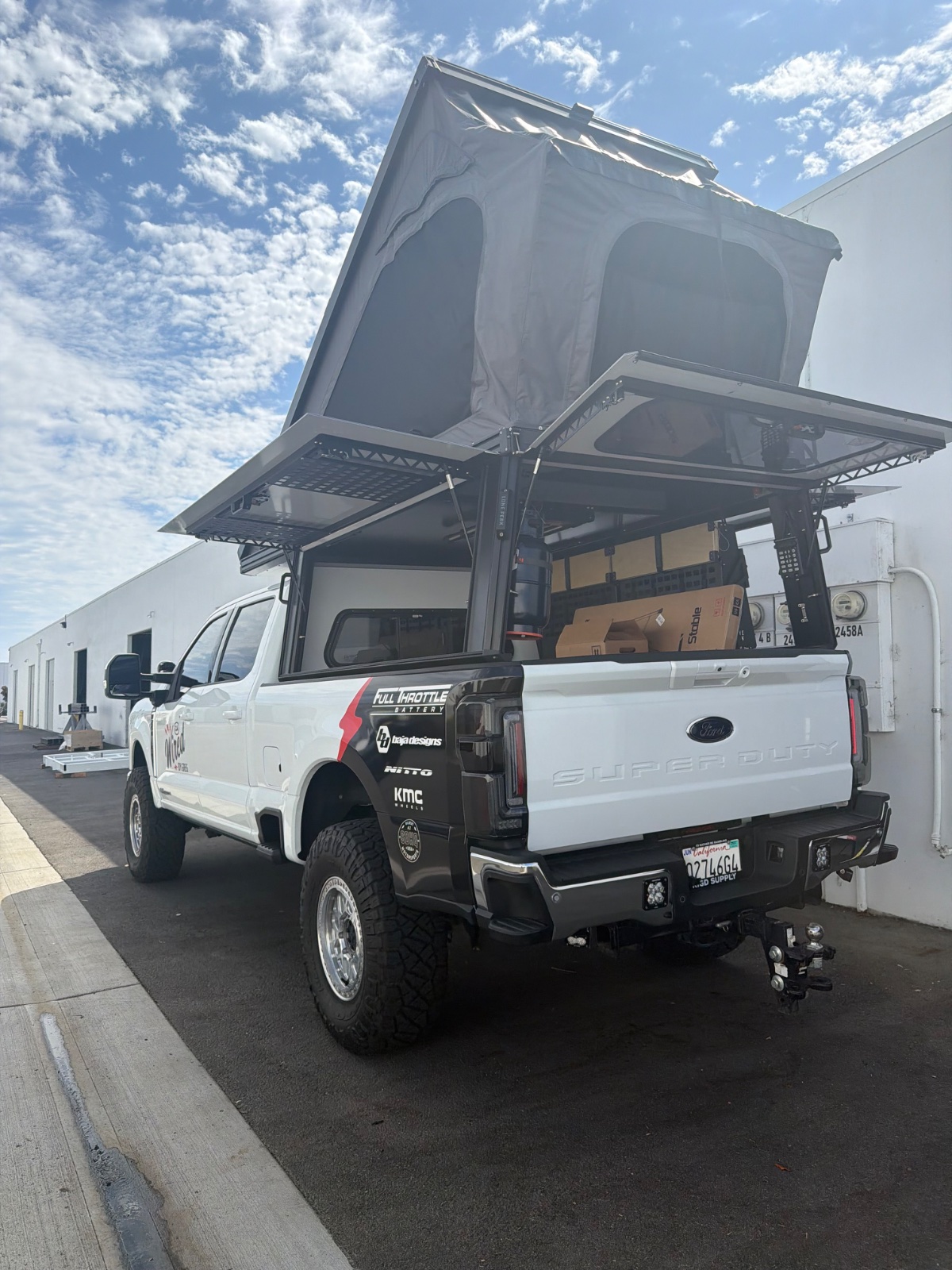 White Ford Super Duty pickup truck with pop-up camper roof open, parked in front of industrial buildings under clear blue sky