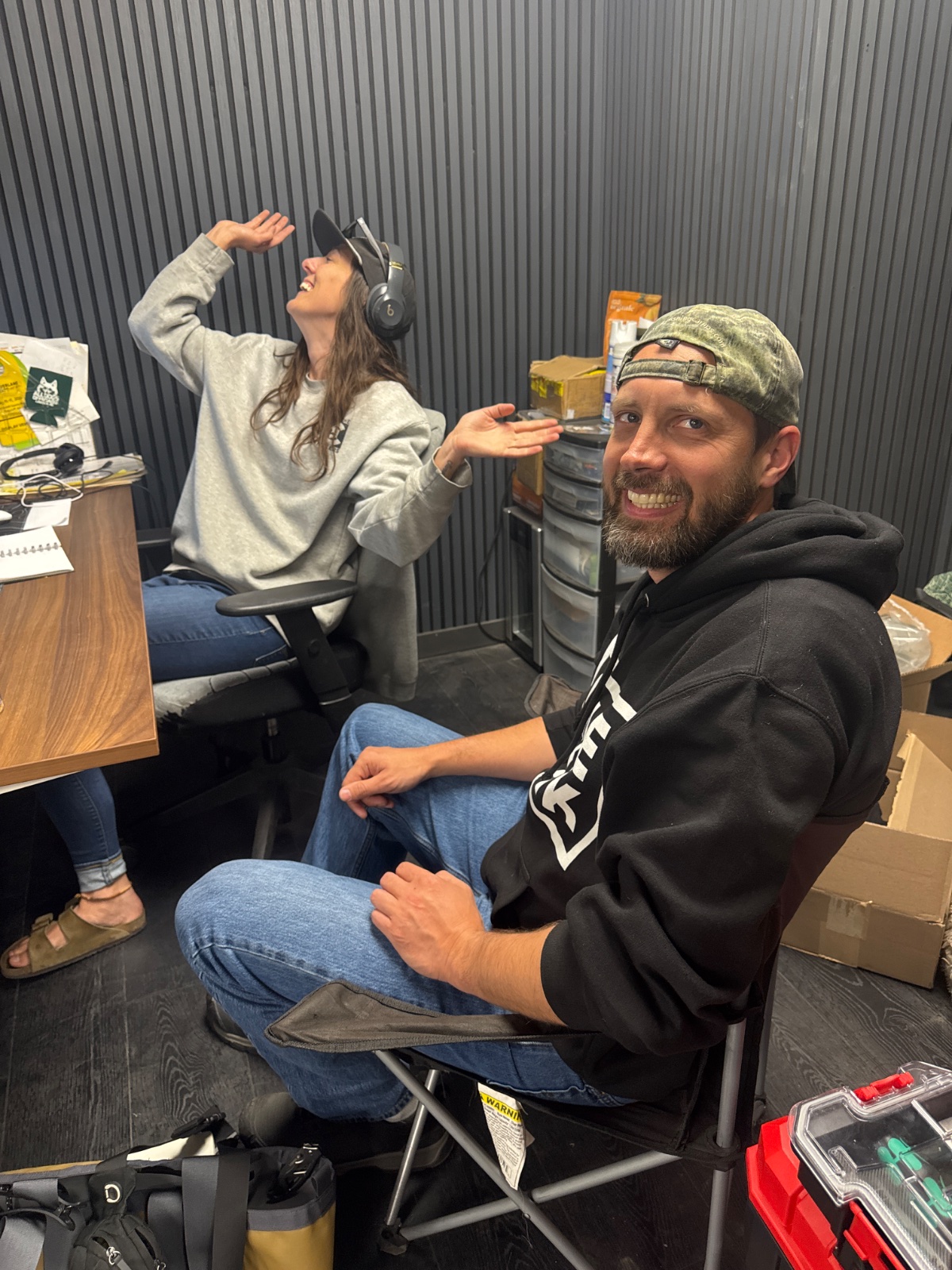 Two people recording a podcast in a metal-walled studio, with the man wearing a black hoodie and cap smiling at the camera while the woman wears headphones and gestures expressively.