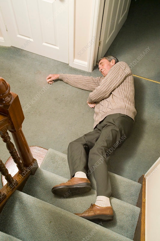 An older man in a tan sweater and dark pants lies on green carpeted stairs, appearing to have fallen, with white doors and wooden railings visible above.