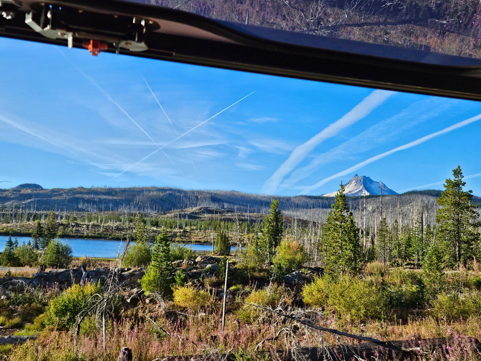 View from an overlook of a volcanic mountain peak, alpine lake, and forest landscape with contrails crossing a blue sky