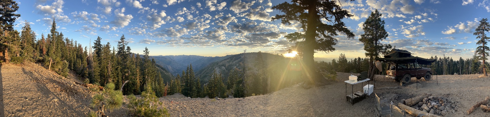 Overlanding camp with black truck and canopy setup on rocky plateau overlooking forested mountain valley at sunset