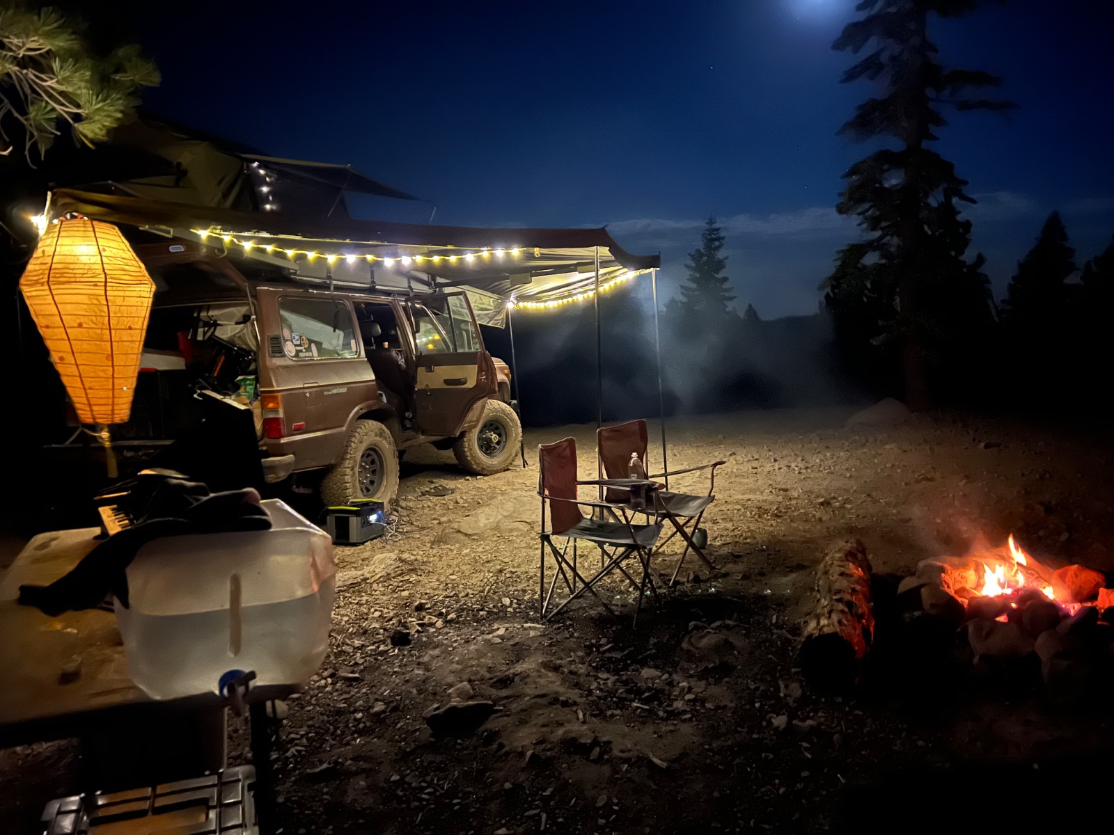 Nighttime overlanding camp with a tan SUV under an awning, campfire, lantern, camp chairs, and evergreen forest backdrop under moonlit sky