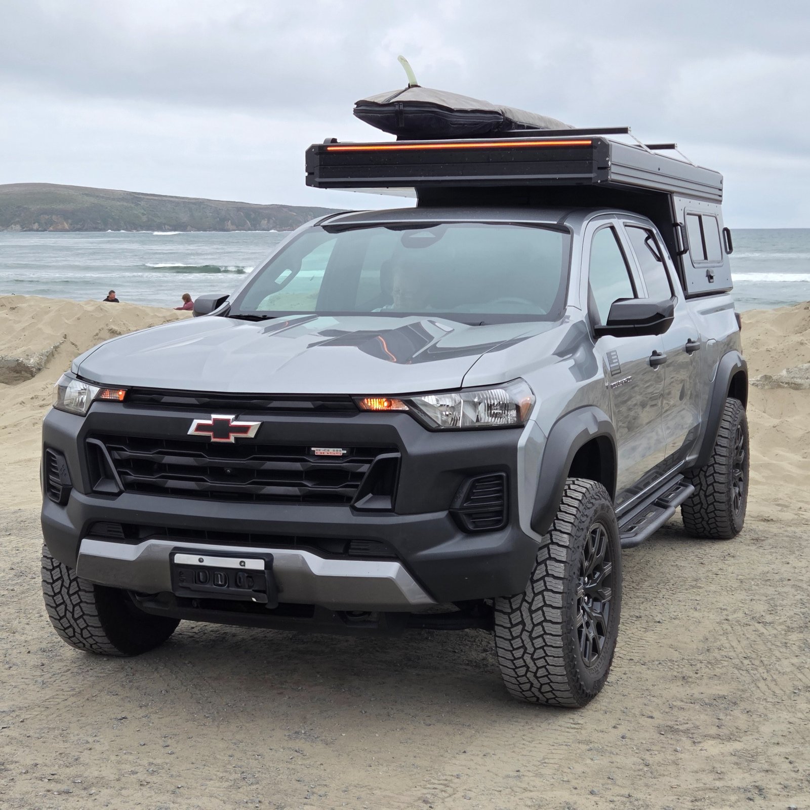Silver Chevrolet Colorado ZR2 pickup truck with roof-mounted camper and kayak parked on sandy beach with ocean and coastal cliffs in background