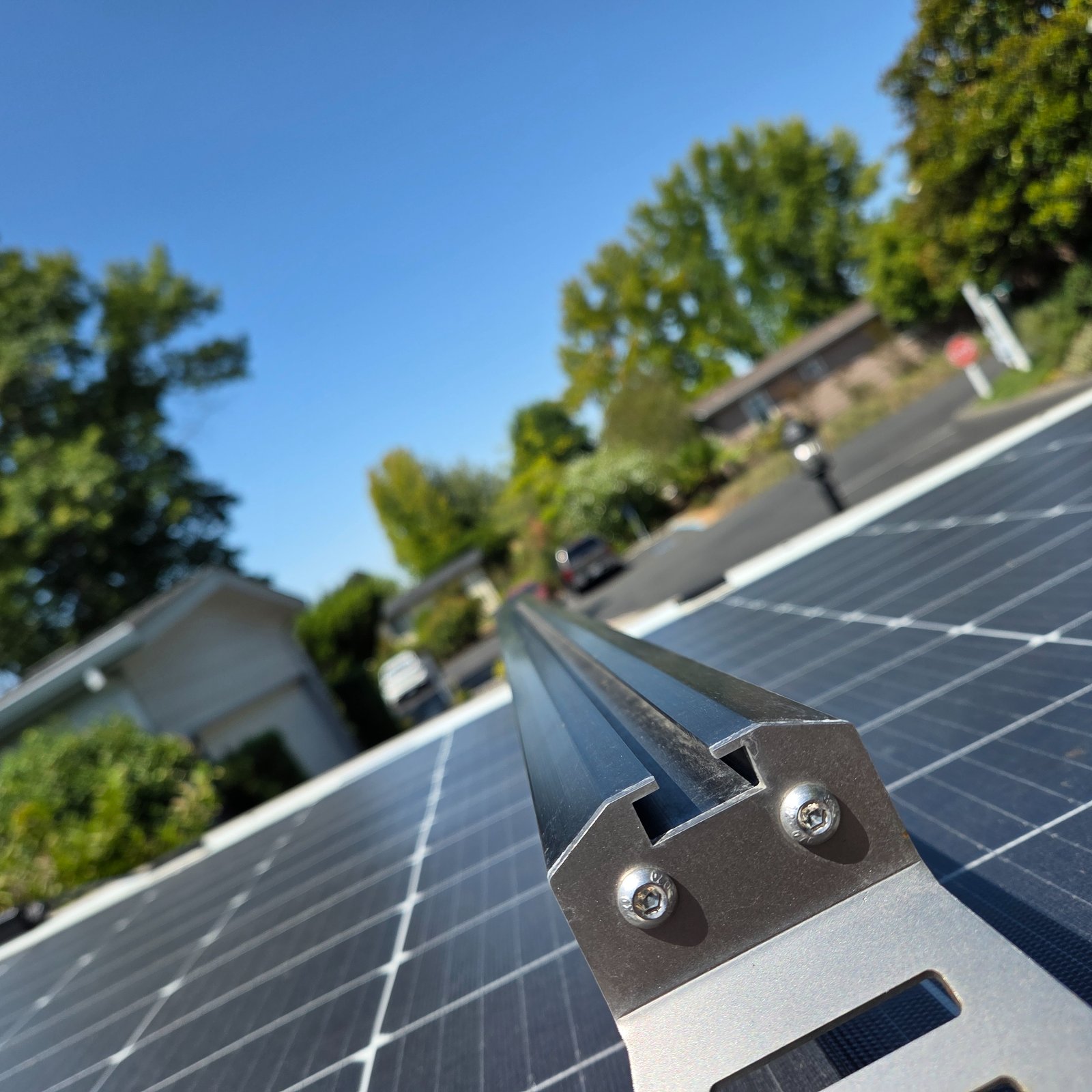 Close-up of a solar panel mounting bracket on a residential rooftop with multiple solar panels installed, surrounded by green trees and suburban houses under clear blue sky