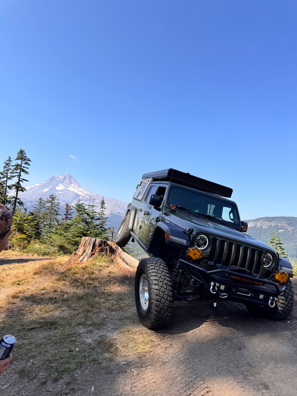 Black Jeep Wrangler with roof rack parked on dirt trail with snow-capped Mount Hood in background, surrounded by coniferous forest