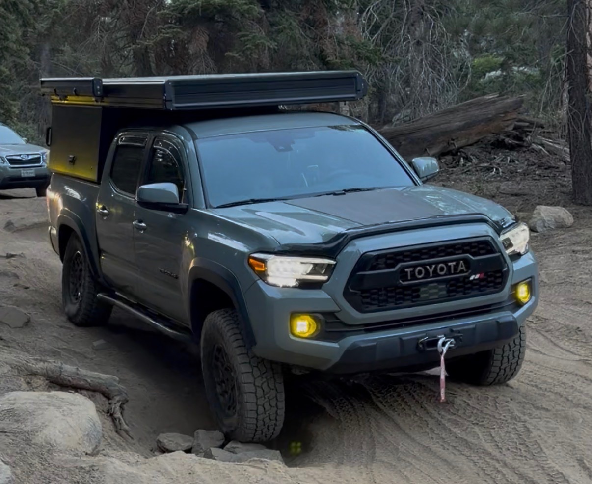 Silver Toyota Tacoma with roof rack and yellow fog lights parked on dirt road near coniferous forest with fallen logs