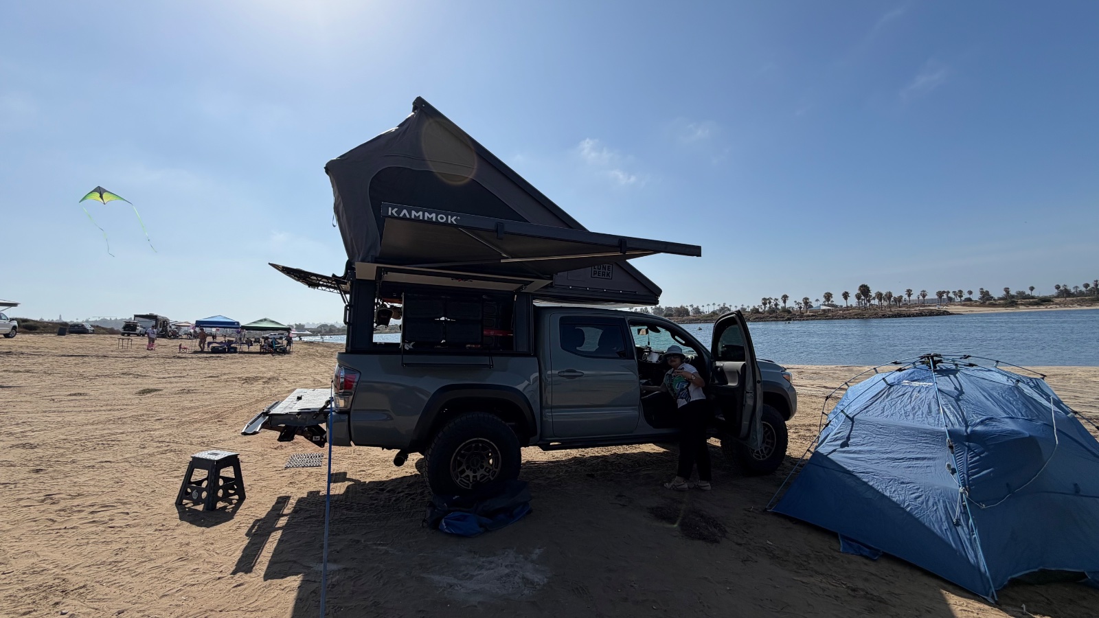 Kammok pop-up camper on a dark pickup truck at a sandy beach with blue umbrella tent, kite flying, and palm trees visible across the water
