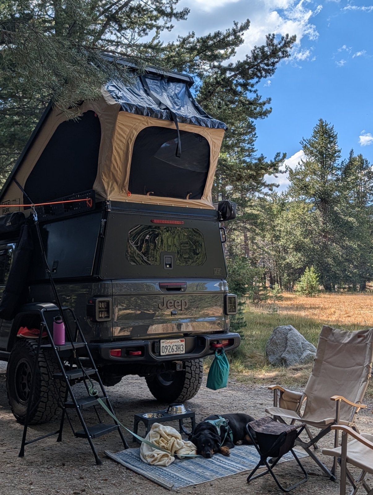 Black Jeep Gladiator with roof-top tent set up at a forested campsite with camping chairs and gear on the ground